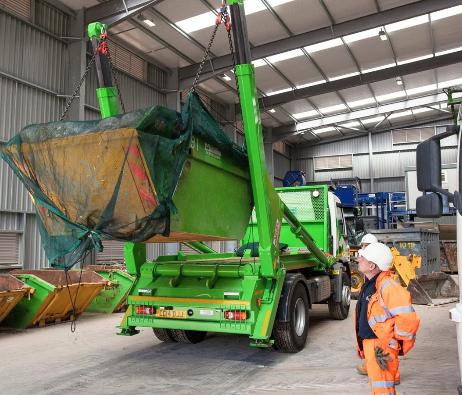 Woodford Recycling skip lorry lifting covered skip container inside UK recycling facility, supervised by worker in high-visibility PPE